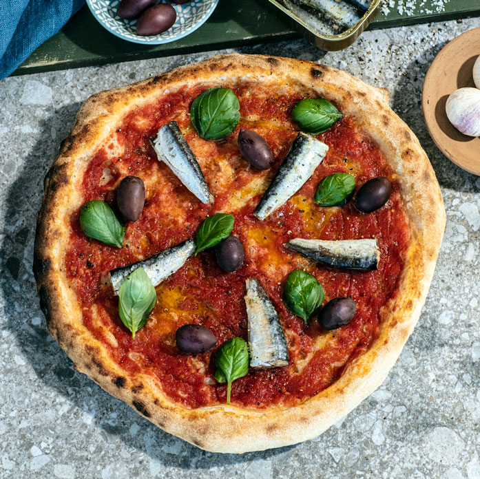 bord med pizza med sardiner, tomatsaus, sorte oliven og basilikum. skål med oliven og hvitløk, boks med sardiner